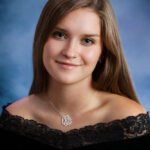 Young woman with long light brown hair in a black lace off-shoulder top and silver necklace, posing for her Senior yearbook portrait against a blue background. "samiseniors.com" watermark is visible.