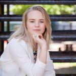 A woman with blonde hair and a white jacket sits on outdoor metal stairs, resting her chin on her hand and looking at the camera—perfect for an outdoor senior portrait or a stylish high school senior yearbook picture.